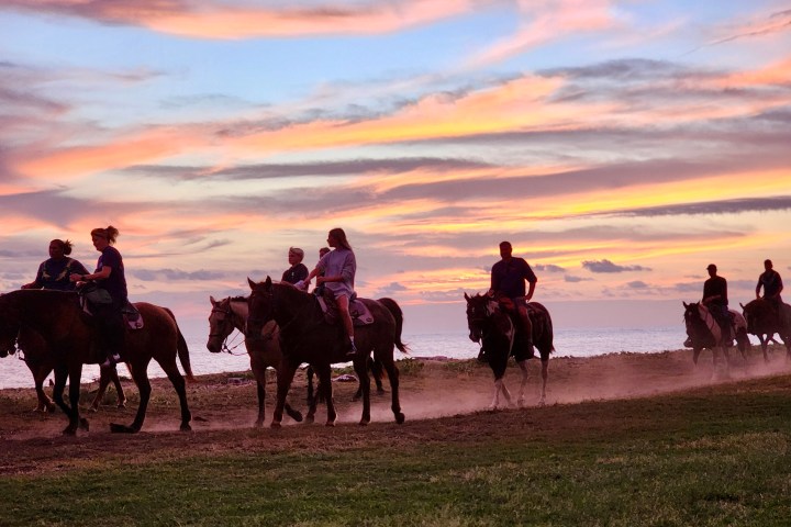 Group of people horseback riding along a beach at sunset with a colorful sky.