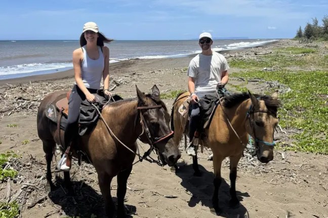 Two people on horseback riding along a sandy beach under a clear blue sky.
