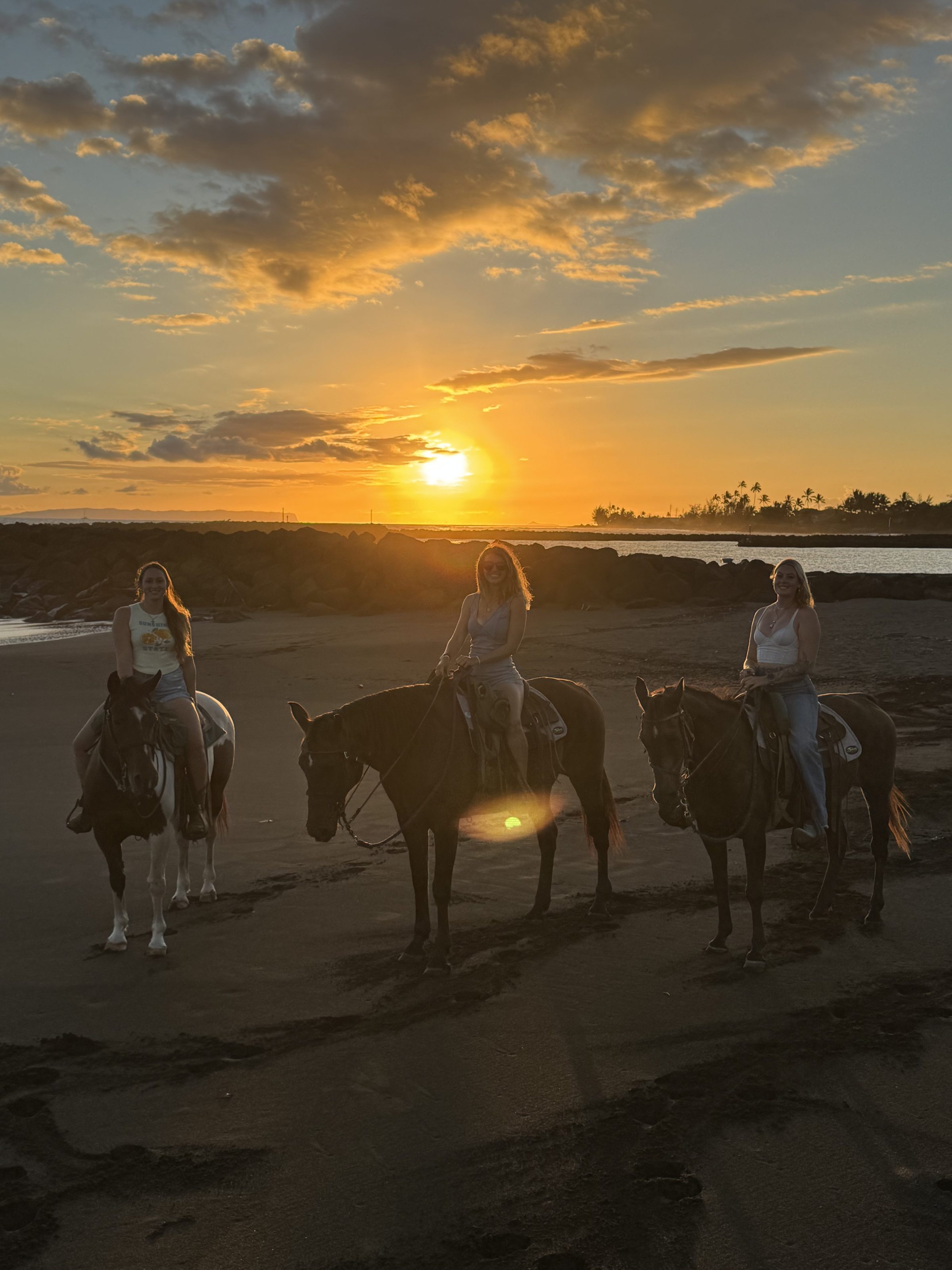 IMG_3448 Three people on horses on a beach at sunset with a vibrant sky.