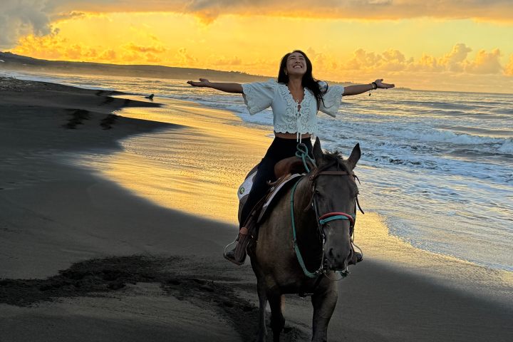 Person riding a horse on a beach at sunset, arms spread wide, with golden waves and dramatic sky.