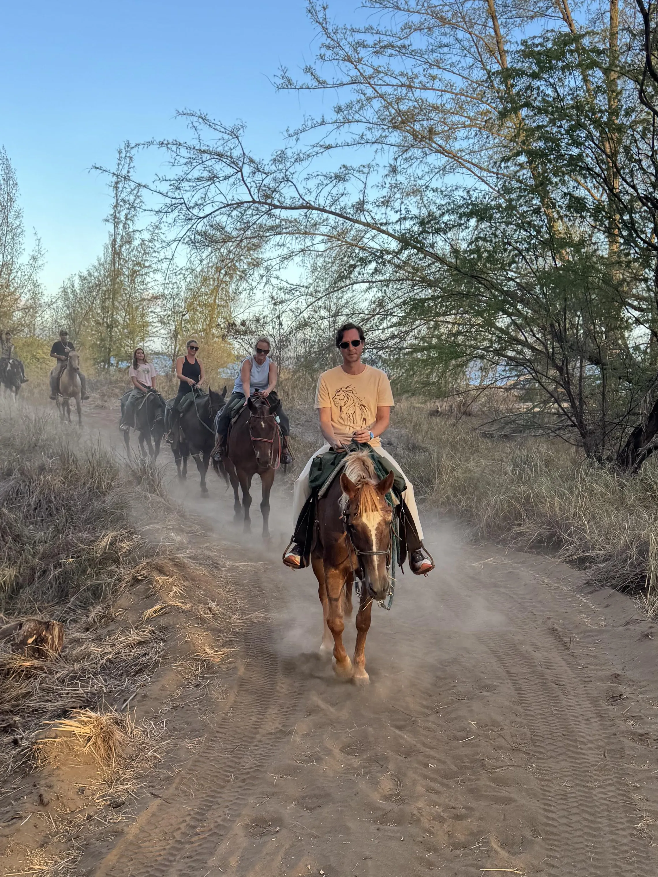 Ranch Group horseback riding on a dirt path surrounded by trees under a clear blue sky.