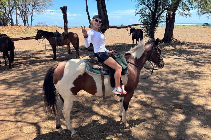 Person on a horse showing a peace sign, under trees, with other horses in the background.