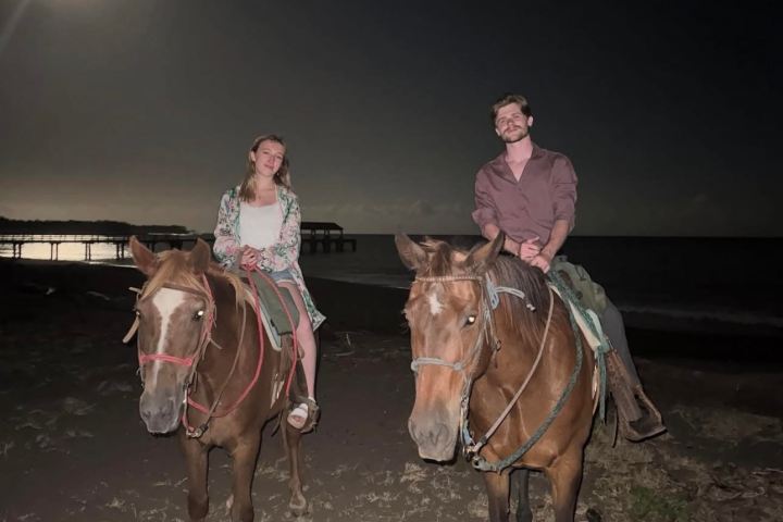 Two people riding horses on a beach at night under a full moon.