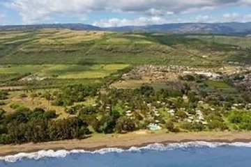 Aerial view of a coastline with a beach, trees, and distant hills under a cloudy sky.